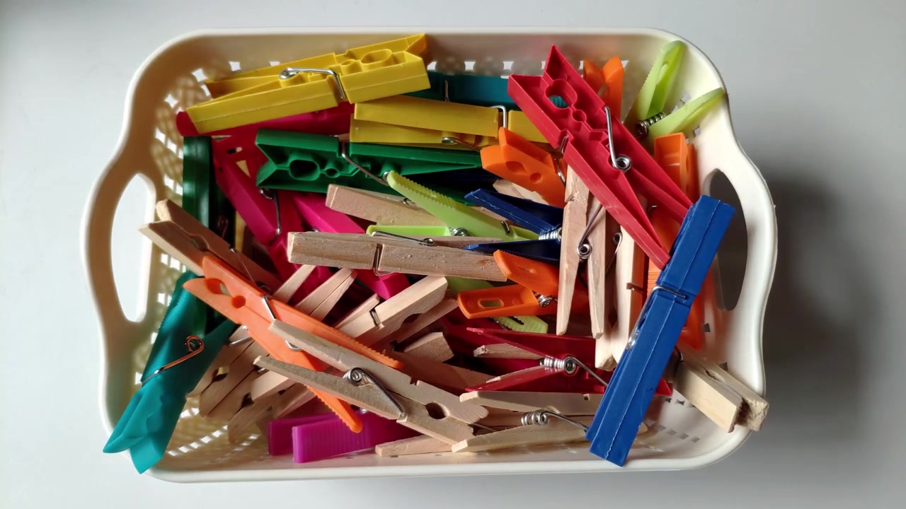 Hånd plukker farverige plastik- og trætøjklemmer fra en kurv på et hvidt bord