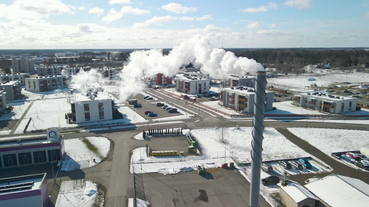 Aerial drone footage spinning around a smoking chimney in Laagri Estonia during a sunny winter day while smoke is coming out of the stack cinemtically and spreads into the blue sky with white clouds.