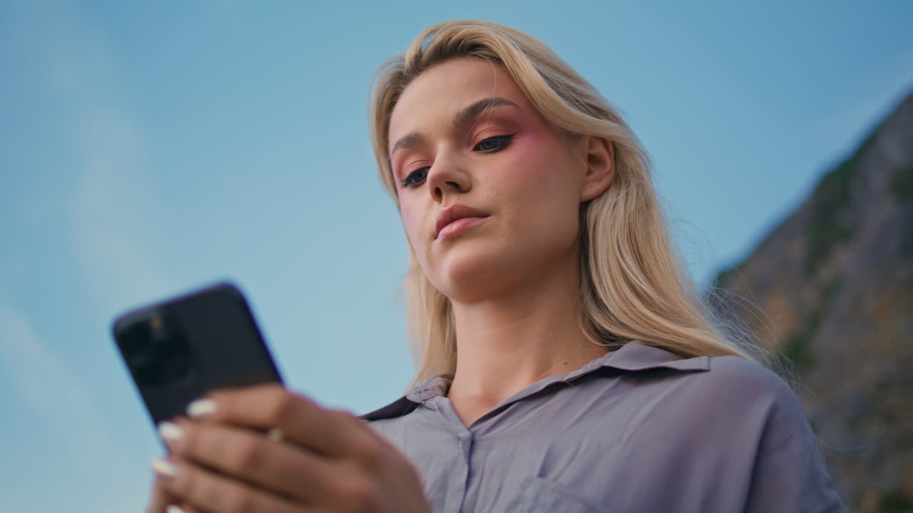 Donna bionda che scrive al cellulare sotto il cielo blu. Donna seria che guarda lo smartphone.
