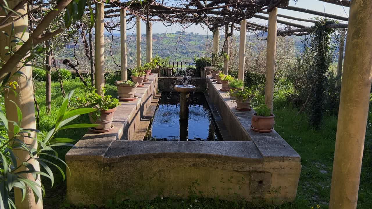 Static shot of a beautiful, ancient stone fountain filled with water, set under a rustic pergola in a green, sunlit garden. Amelia, Umbria, Italy