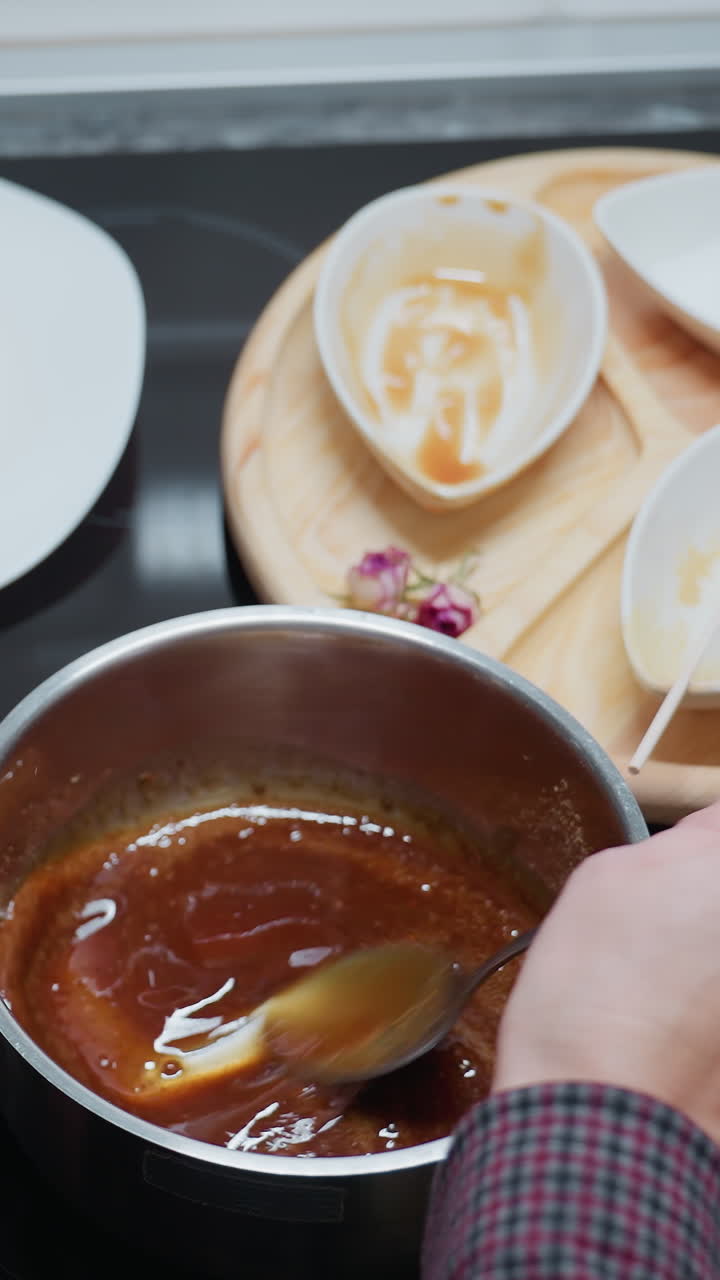 vista parcial de una persona mezclando sopa en una olla de acero inoxidable con una cuchara, ingredientes mezclándose en líquido, bandeja de madera con cuencos y utensilios usados en la encimera de la cocina de mármol oscuro
