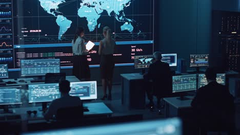 two software engineers talking next to a big screen in a modern monitoring office with live analysis feed with charts. monitoring room big data scientists and managers sit in front of computers.