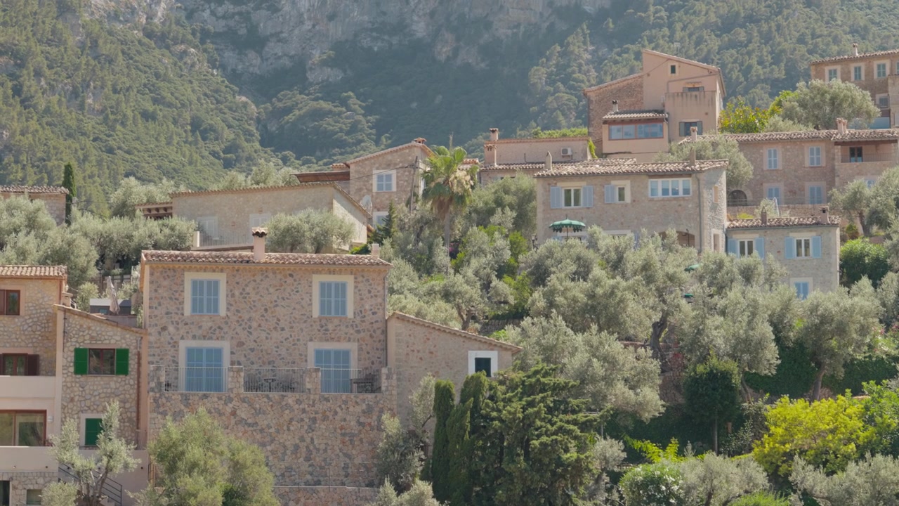 Ville rustiche in pietra tra gli ulivi sui pendii terrazzati di Deia, con una montagna boscosa che si erge sullo sfondo. Calda luce estiva mediterranea. Inquadratura ampia.