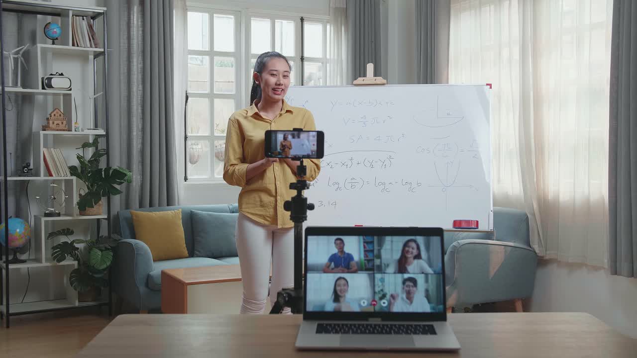 Asian Female Teacher Shoots Video By Smartphone Camera And Having Video Call On Laptop While Teaching Math At Home