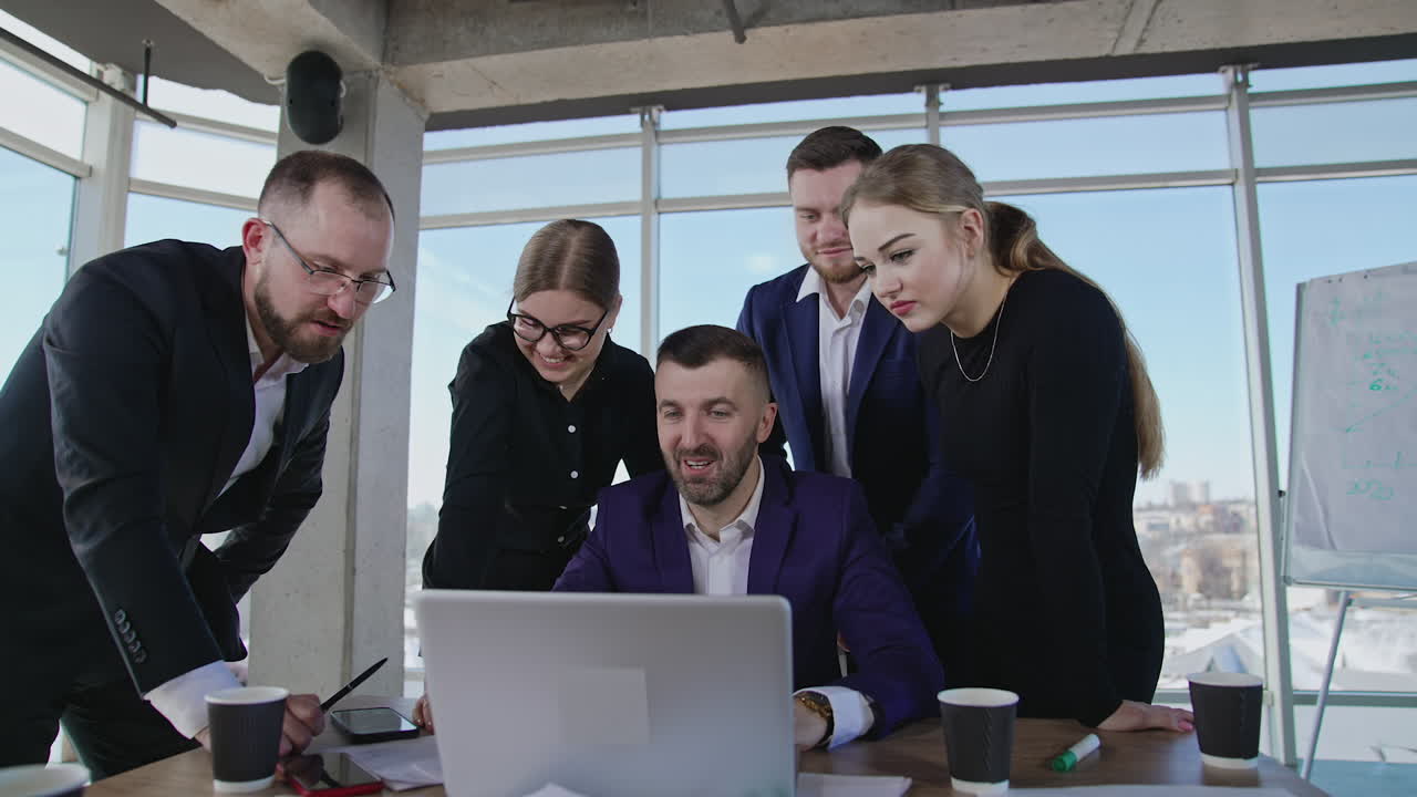 Business team standing around the chief and looking at laptop. One of the male colleagues showing his smartphone.