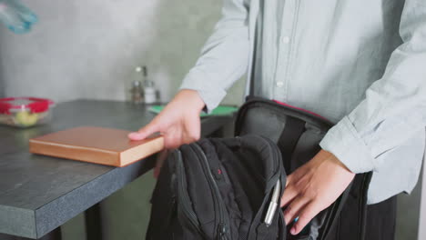 boy in light shirt packing school bag on table with water bottles, food containers, and books nearby, calmly preparing for school day in kitchen setting with organized items and focused motion