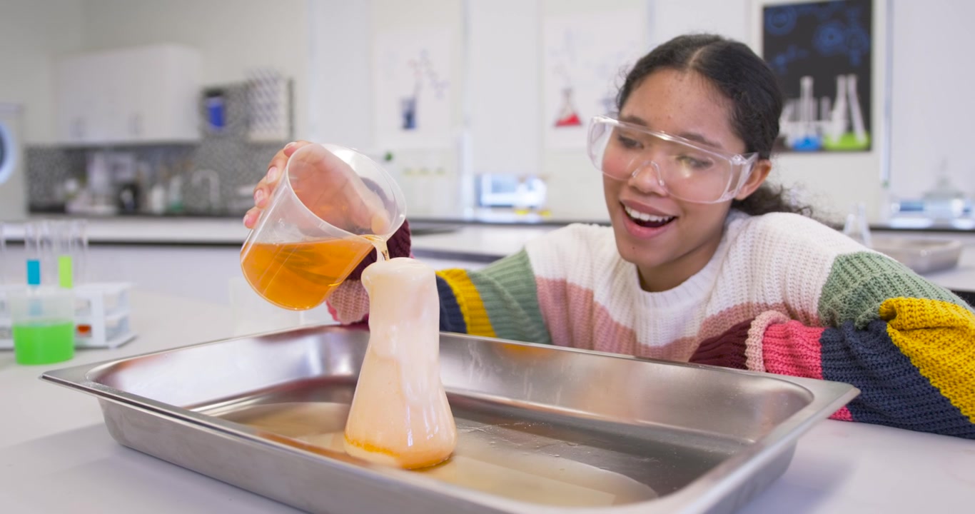 Niño vertiendo líquido naranja de un vaso de precipitados a un matraz en una demostración de laboratorio, causando un desbordamiento de espuma, sonriendo, con espacio para texto