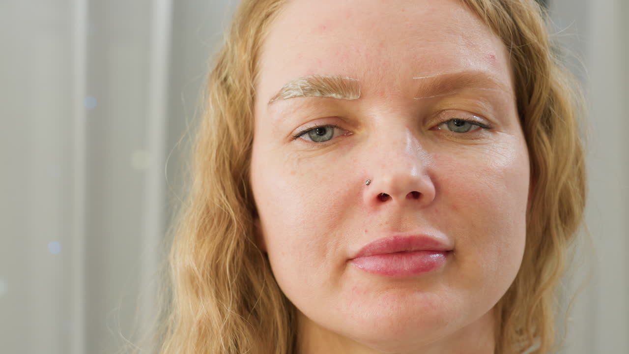 Close-up of client wearing nose ring during beauty session with visible cream applied on one eyebrow while the other remains untouched, showing contrast in eyebrow treatment process in clean salon