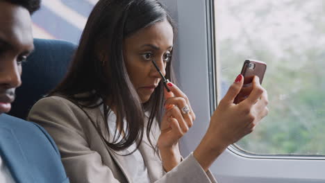 businesswoman sitting in train commuting to work putting on make up using mobile phone as mirror