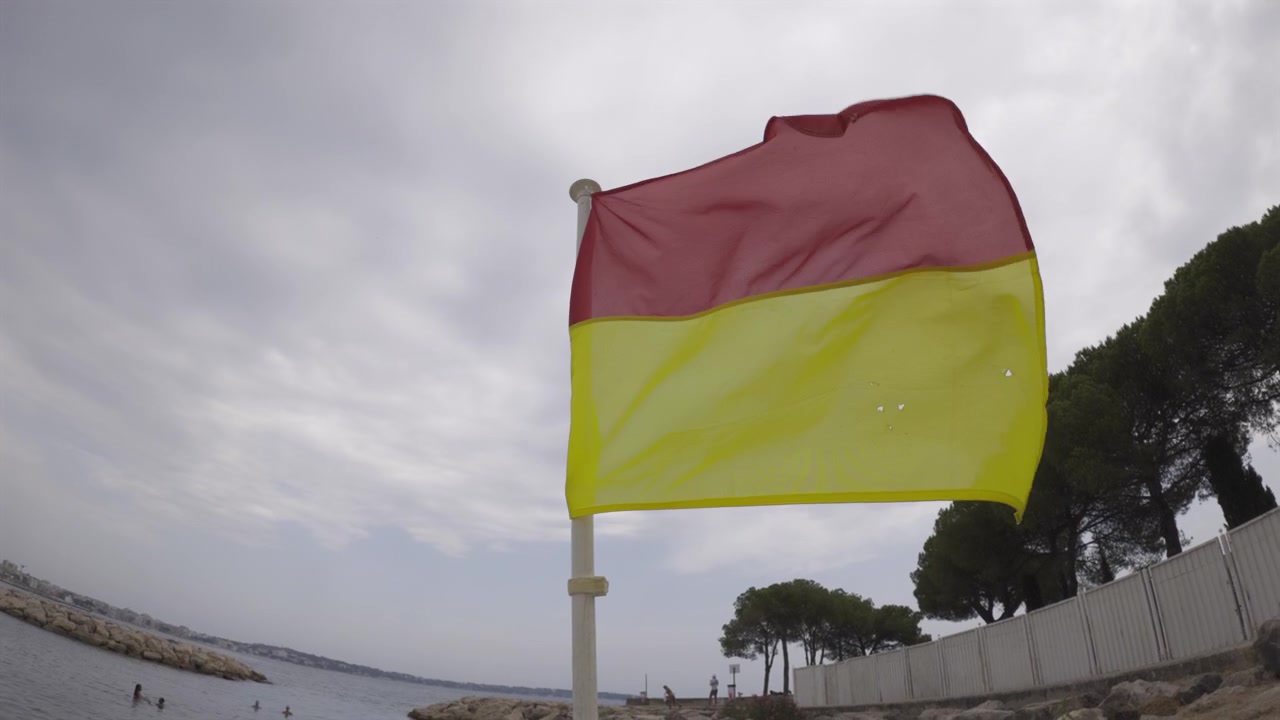 Bandiera rossa e gialla che sventola sulla spiaggia, segnalando una zona di balneazione sicura con acqua blu calma e una costa rocciosa sotto un cielo nuvoloso.