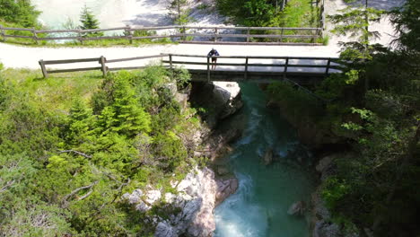 man-standing-on-bridge-over-fast-river-rapids-in-the-forest-of-Italian-Dolomites-during-summer,-aerial