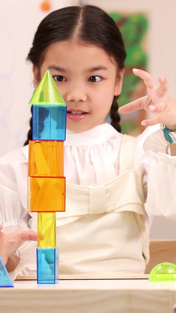 Elementary girl carefully stacks colorful magnetic blocks into tall tower during bright classroom STEM lesson
