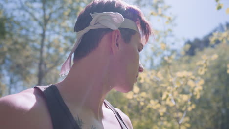 side view of adventurer bending to tie headband under freckled hair while remaining vigilant under dazzling sunlight in forest clearing, arms raised, sweat glistening, expression determined