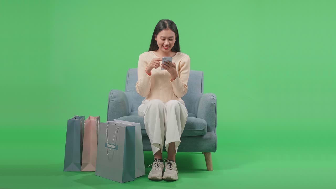 Front View Of An Asian Shopping Woman With Shopping Bags Enjoy Using Mobile Phone Shopping Online While Sitting On Sofa In Front Of Green Screen