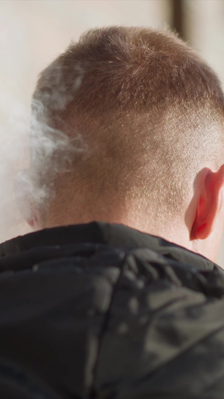 Close up of man head and neck from behind showing short hair and ears with slight movement. subject wears black jacket while standing outdoors in bright light smoke appears briefly near top