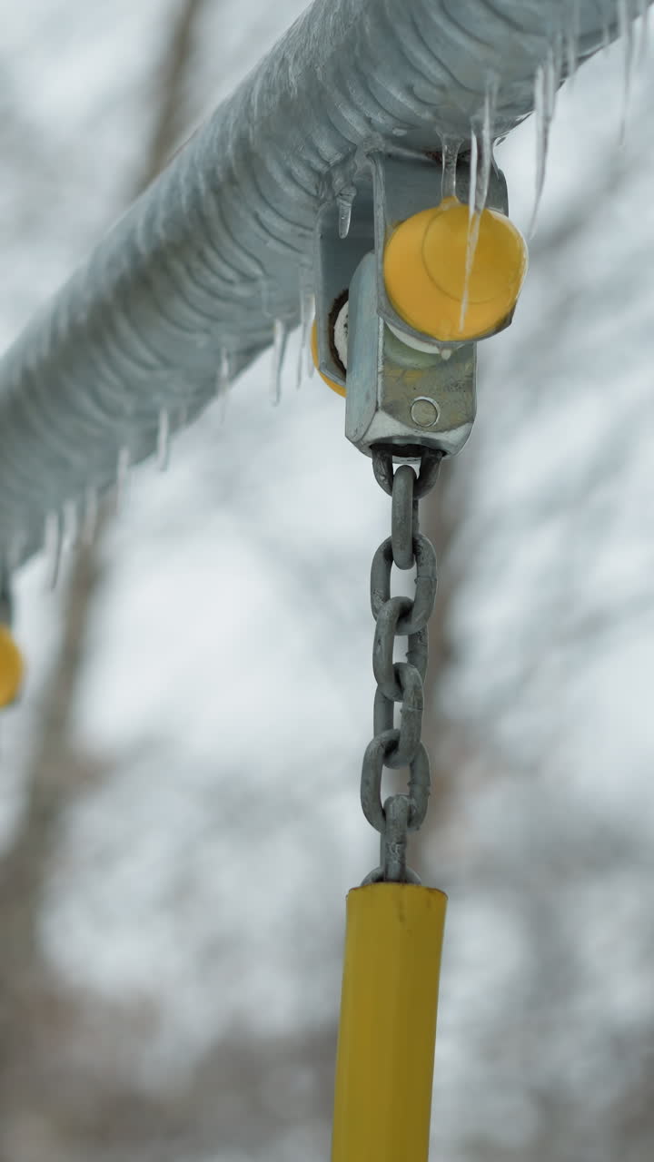 primer plano de una articulación metálica de cadena oscilante con tapón amarillo cubierto de hielo y hielo colgando a lo largo de una barra de metal congelada, en un fondo invernal borroso con árboles desnudos y una atmósfera serena