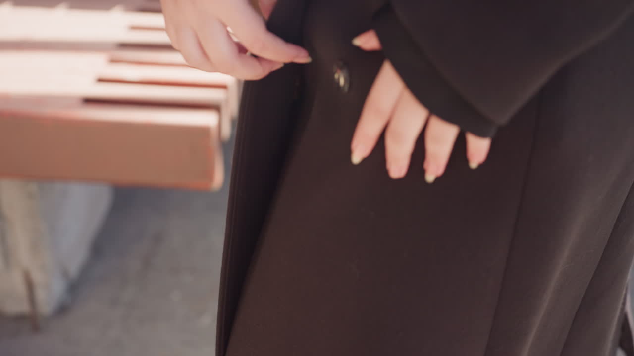 어두운 겨울 코트의 스냅 버튼을 클로즈업해서 보여주고 손이 스쳐 지나가는 모습이에요. 햇빛이 벽돌 배경 근처의 울 텍스처를 비추고, 외장재 하드웨어와 미니멀한 도시 패션의 순간을 강조하는 장면이에요.