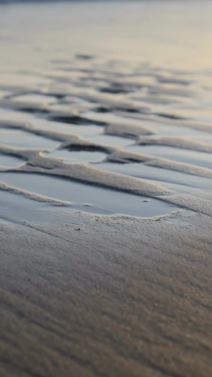 Vertical video: Retreating tide pulling foam line exposing wet ridged sand at shore, shallow pools