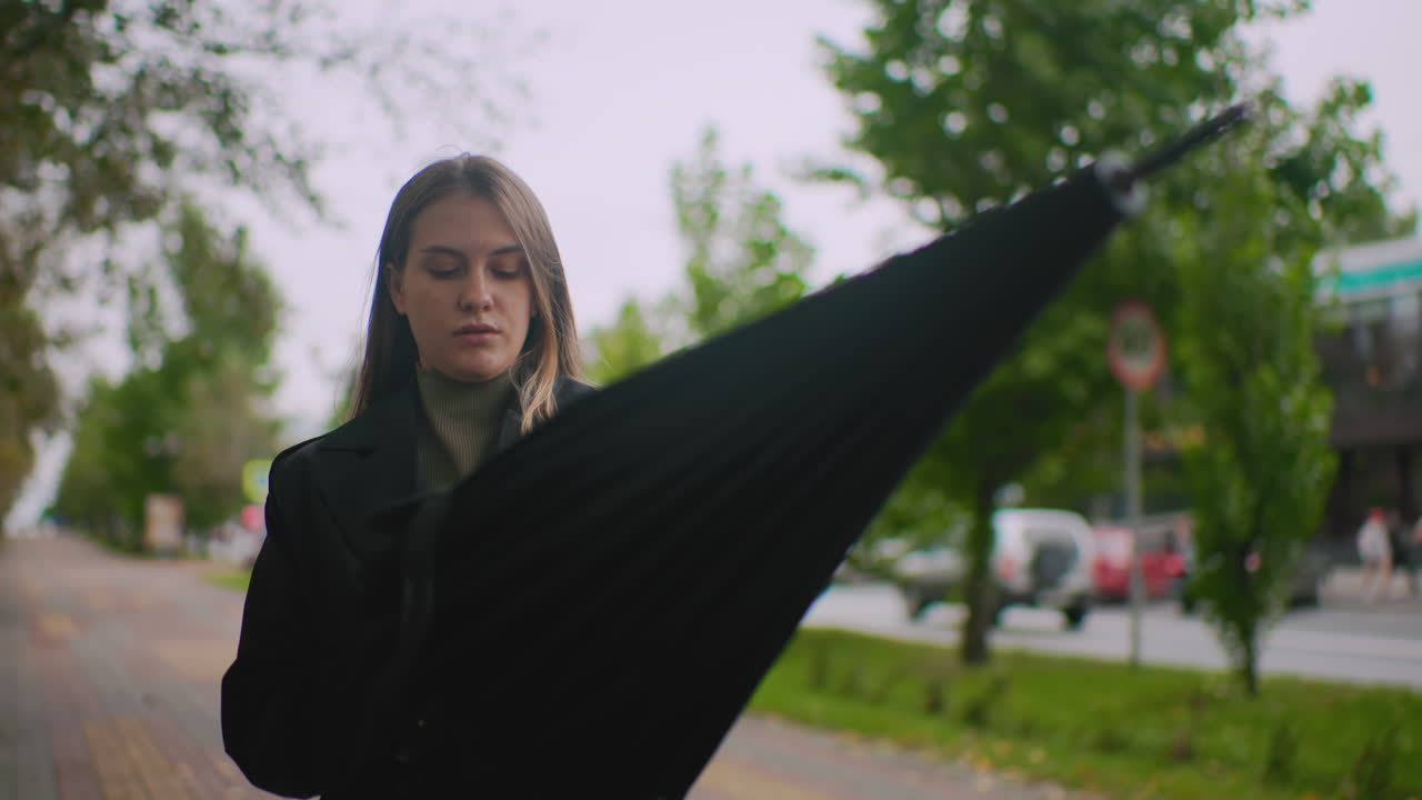 Woman in black coat stands outdoors looking up at sky with raised hand, anticipating rain, holding umbrella handle, urban park background with greenery and blurred buildings, capturing thoughtful expression
