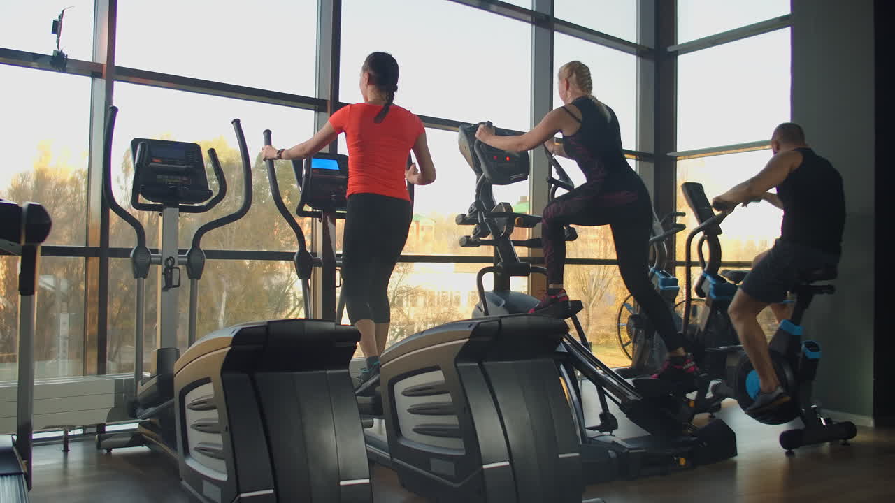 Young fit woman using an elliptic trainer in a fitness center. A group of young women train on sports training equipment in a fitness gym. Steady cam shot.