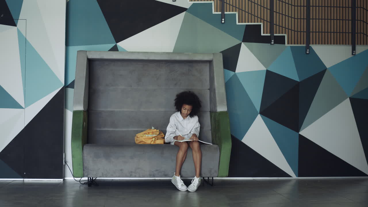 African american schoolgirl drawing in school corridor. Curly girl sitting bench