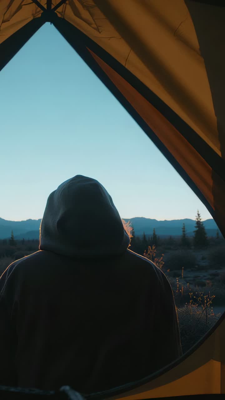 Video verticale: persona in piedi con felpa con cappuccio rivolta verso la linea di cresta nell'apertura della tenda, mentre l'alba si fa più luminosa