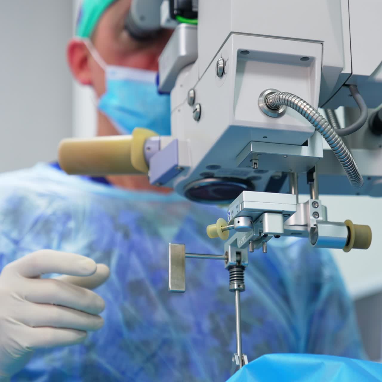 Adult male surgeon in mask and gloves sits behind the equipment. Doctor looks into binoculars attentively then turns to take instrument