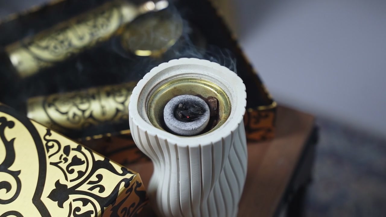 Primo piano dall'alto di carbone ardente che brucia bakhoor all'interno di un bruciatore in ceramica bianca con fumo che sale accanto a scatole di lusso con motivi dorati.