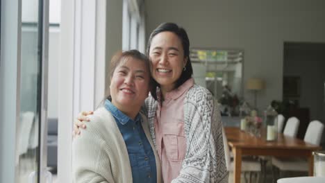 portrait of happy asian mother and adult daughter embracing and smiling in dining room