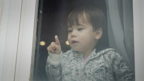 a two-year-old azait boy looks out of the window, emotionally points his finger down