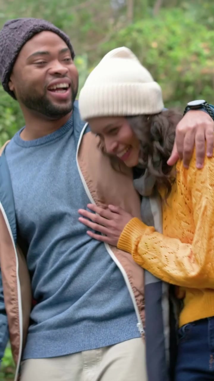 Video verticale: Uomo e donna sorridenti camminano su un sentiero del parco, il contatto visivo sfocia in un tocco giocoso, entrambi con berretti.