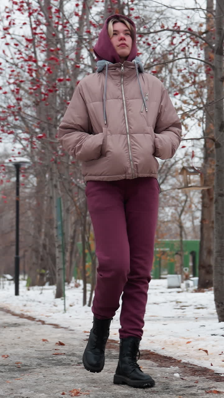 Una donna con un caldo abbigliamento invernale cammina con le mani in tasca su una strada innevata fiancheggiata da alberi spogli adorni di bacche rosse, con impronte visibili sul sentiero ghiacciato.