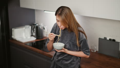 young house wife with long flowing hair eating long pasta with chopsticks while resting casually against kitchen cabinet in bright modern kitchen with blurred kitchen equipment in soft background
