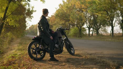 Ein-Junger-Attraktiver-Mann-In-Lederjacke-Und-Stylischer-Sonnenbrille-Sitzt-An-Einem-Sonnigen-Herbsttag-Auf-Seinem-Fahrrad-Auf-Der-Landstraße