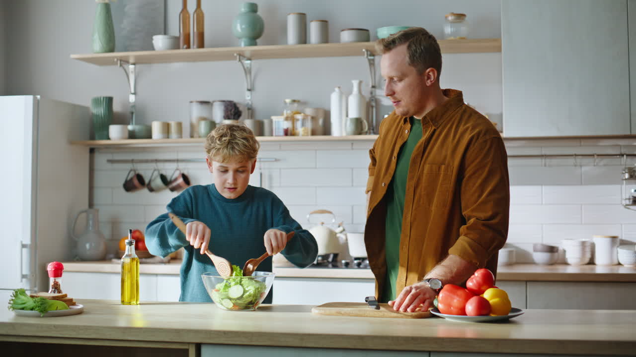 Nahaufnahme: Kleiner Sohn gibt Öl zu Salat, kocht mit Vater Frühstück in der Küche.