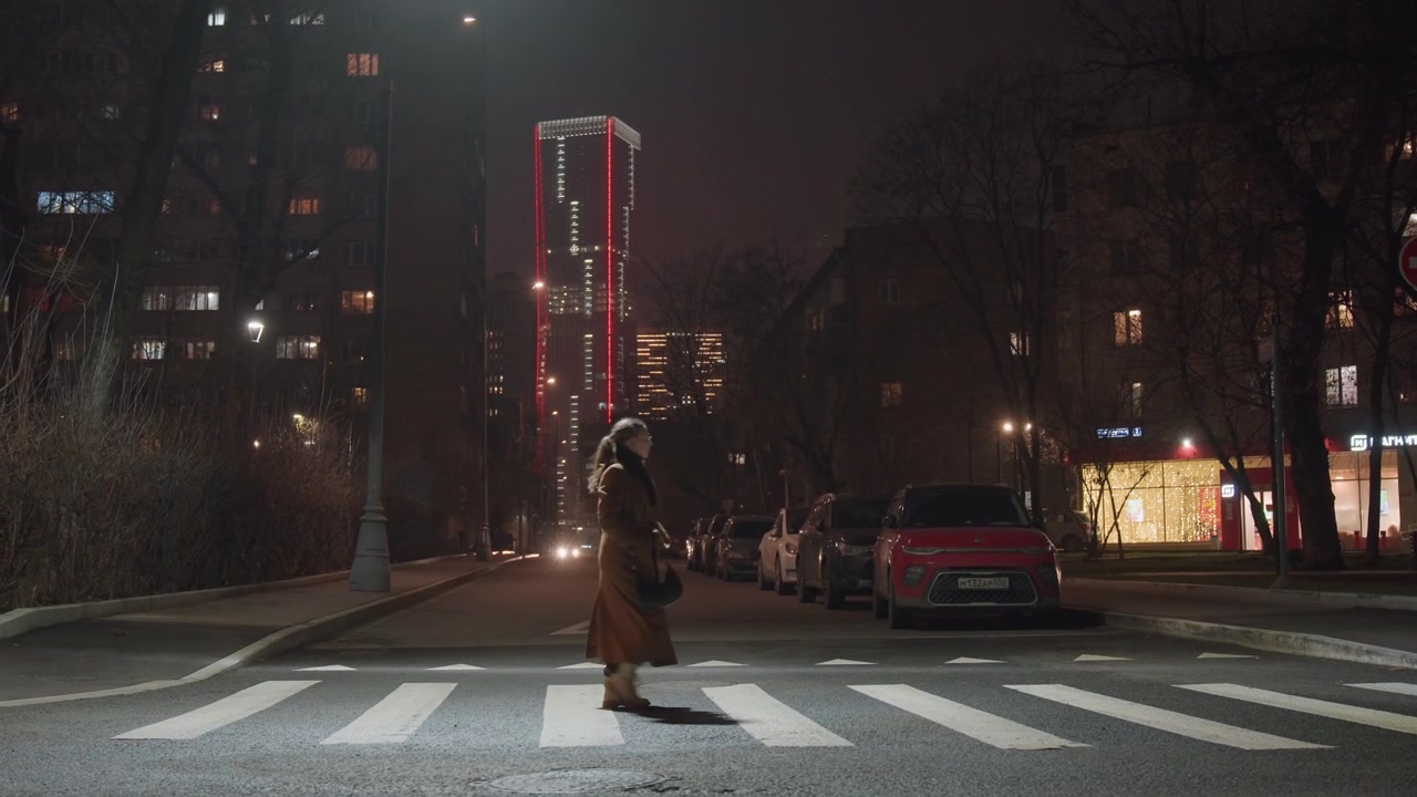 Night crosswalk under neon tower, lone pedestrian crossing zebrastriped street, parked car with glowing headlights, lit storefronts and redlit skyscraper in distance, falling snow on sidewalks