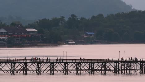 Mon-Bridge-and-a-longboat-followed-speeding-behind-the-structure-while-people-are-walking-on-the-bridge,-silhouetting-as-it-was-getting-dark,-in-slow-motion