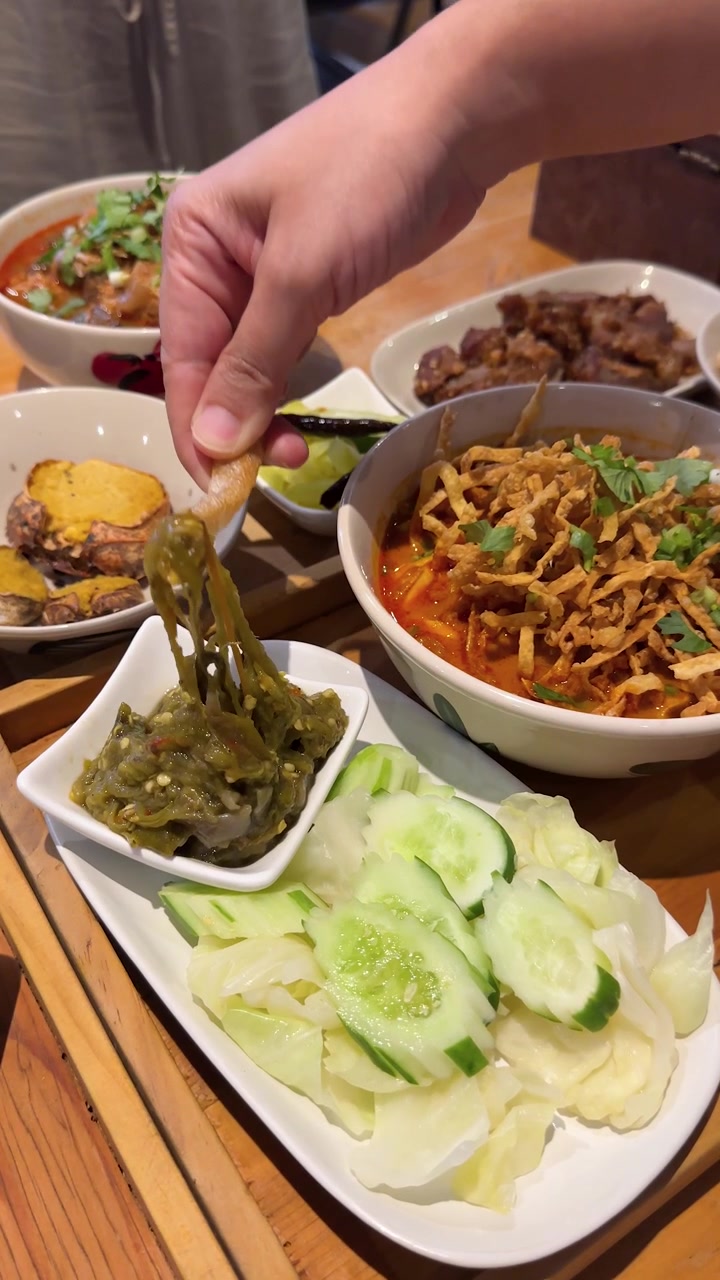 มือจุ่มแคบหมูกรอบลงในน้ำพริกหนุ่มข้างแกงข้าวซอย