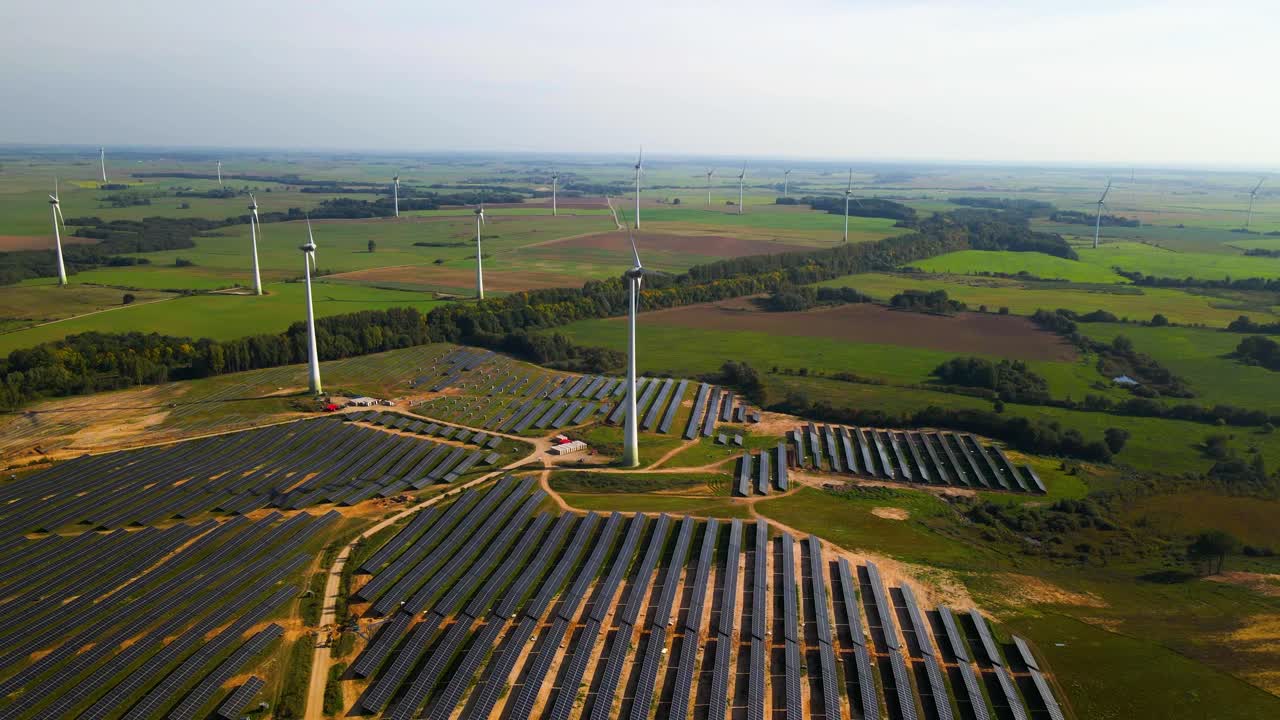 Aerial footage of solar panels plant and wind turbines in a wind farm generating green electric energy on a wide green field on a sunny day, in Taurage, Lithuania, zooming in