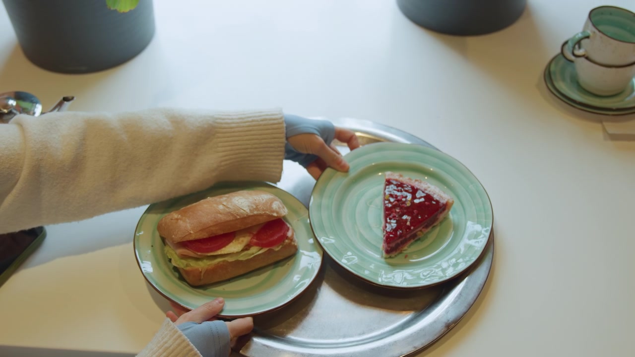 Primo piano di una persona che sistema alimenti su un vassoio d'argento. Una mano posiziona costantemente piatti verdi con un sandwich e una torta sul tavolo. L'ambiente è uno spazio interno luminoso con pareti bianche.
