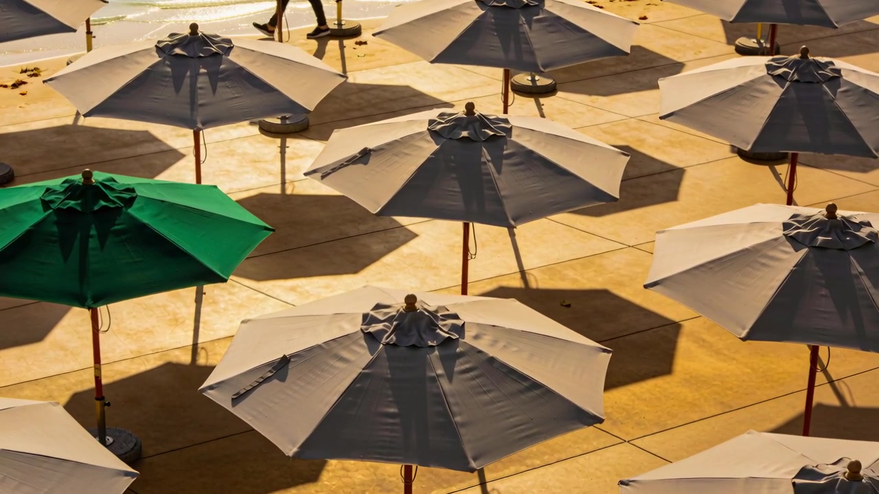 Champ de parasols sur le pont, parasol vert, la caméra zoome tandis qu'un homme en vêtements sombres marche sur le rivage.
