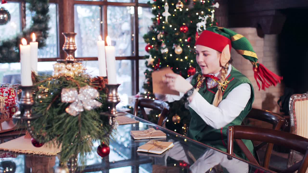 Cheerful elf girl sitting at a wooden table looking through Santa Claus mail. Room with Christmas decorations