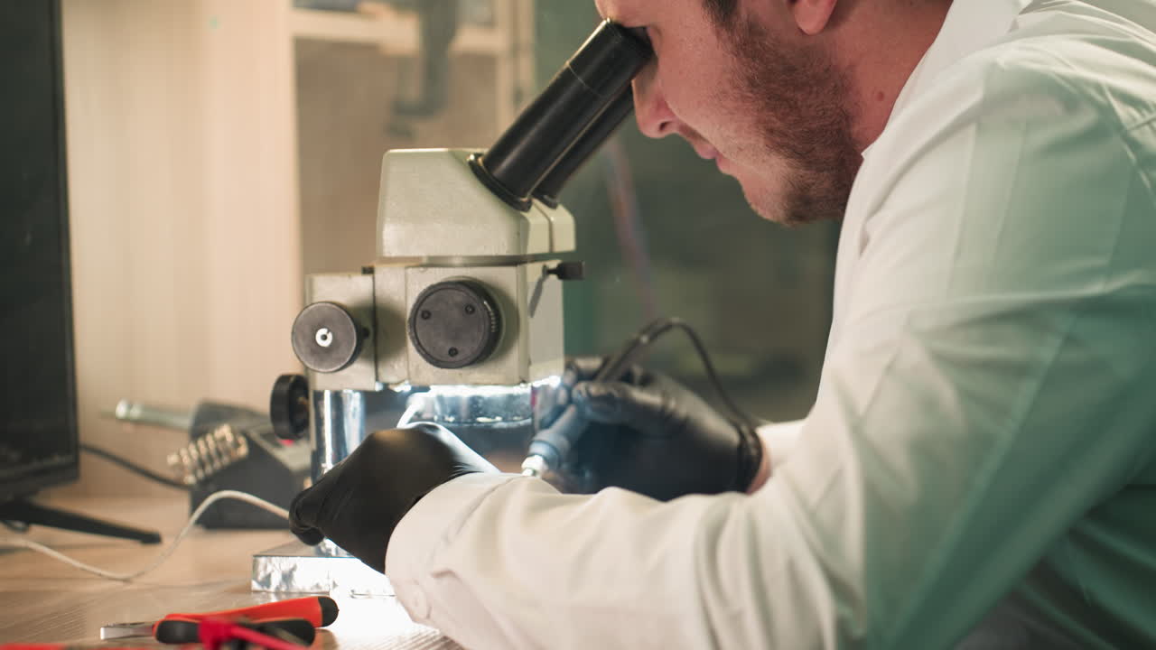 una vista de cerca de un técnico enfocado en una bata de laboratorio trabajando cuidadosamente en una placa de circuitos electrónicos bajo un microscopio