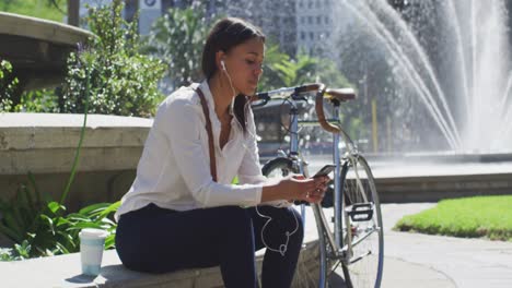 african american woman with bike using smartphone wearing earphones in city park