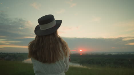 close-up back view of a woman in a white dress, focusing on her hair and black hat, as she stands alone in a serene grassy field, arms folded, gazing over a tranquil lake during a peaceful sunset