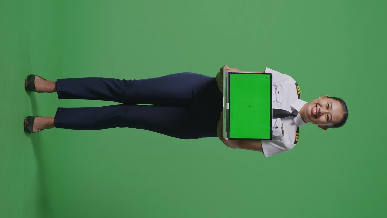 cuerpo lleno de piloto mujer asiática sonriendo y mostrando pantalla verde portátil a la cámara mientras estaba de pie en el estudio de fondo pantalla verde