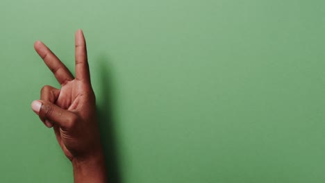 Close-up-of-hand-of-biracial-man-showing-peace-sign-with-copy-space-on-green-background