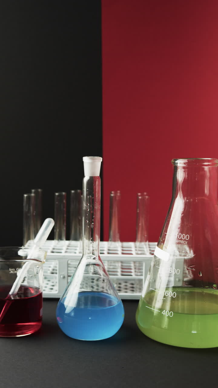 Vertical video of coloured liquids in flasks with test tubes in stand on black and red background