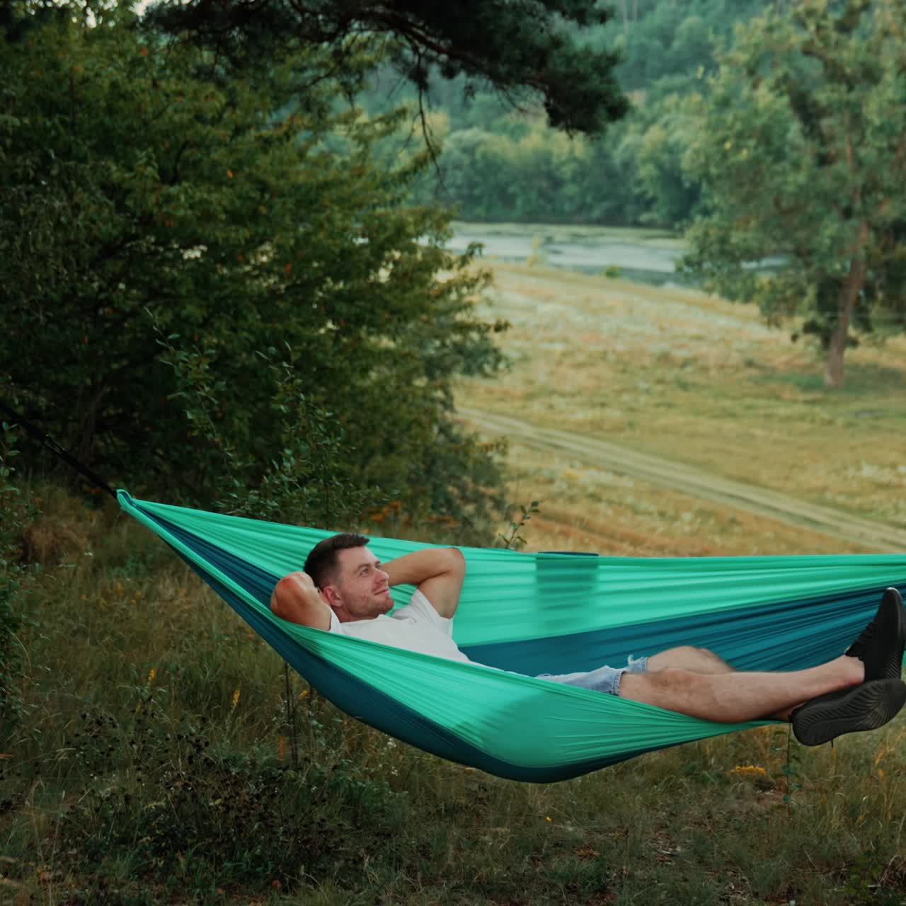 Happy Caucasian man lies comfortably in a hammock hands behind his head. Relaxed person looks dreamily up swaying slowly in hammock. Nature at backdrop