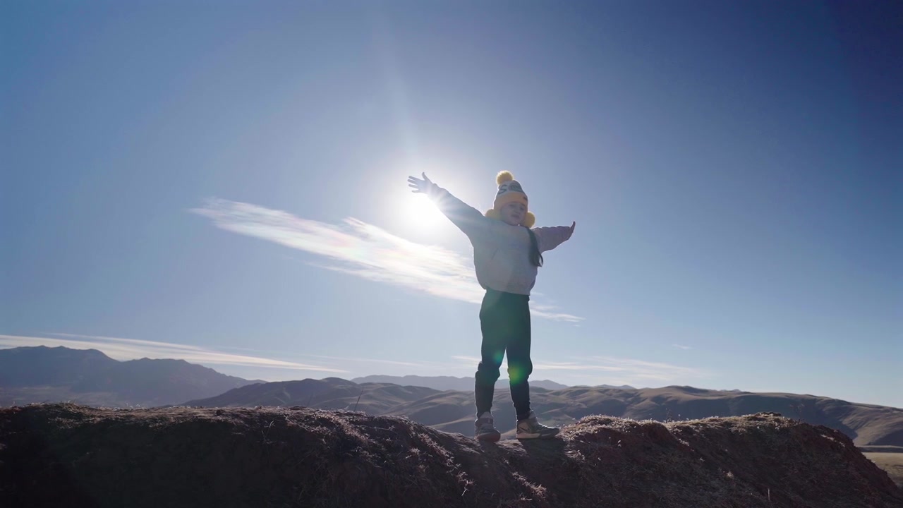 Young girl stands with arms outstretched on a rocky ridge in Tafí del Valle, Tucumán, Argentina, backlit by the sun during a family adventure, slow motion camera push in.